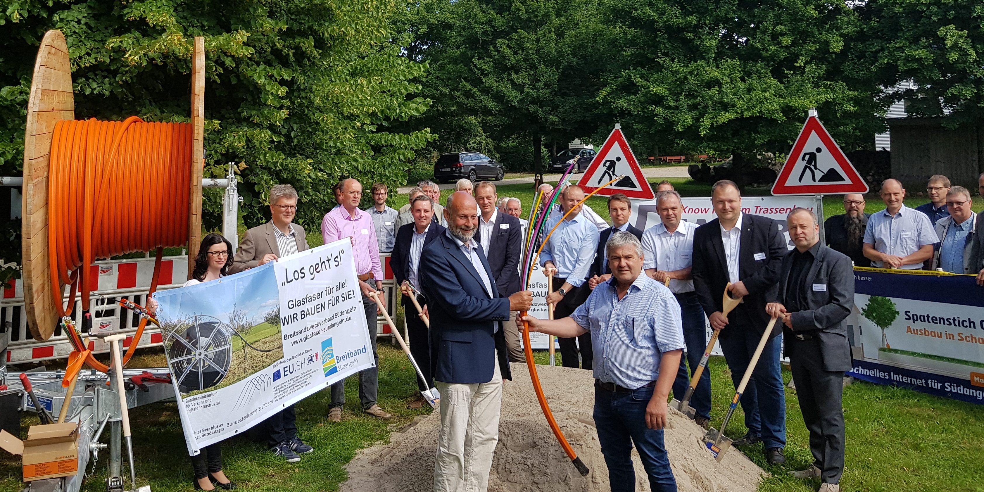 Eine Gruppe von Menschen steht auf einer grünen Wiese. Zwei Männer halten ein orangefarbenes Kabel in der Mitte, umgeben von Schildern und Geräten. Im Hintergrund sind Bäume und mehr Personen.