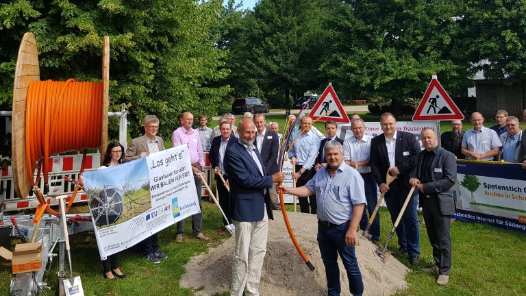Eine Gruppe von Menschen steht auf einer grünen Wiese. Zwei Männer halten ein orangefarbenes Kabel in der Mitte, umgeben von Schildern und Geräten. Im Hintergrund sind Bäume und mehr Personen.