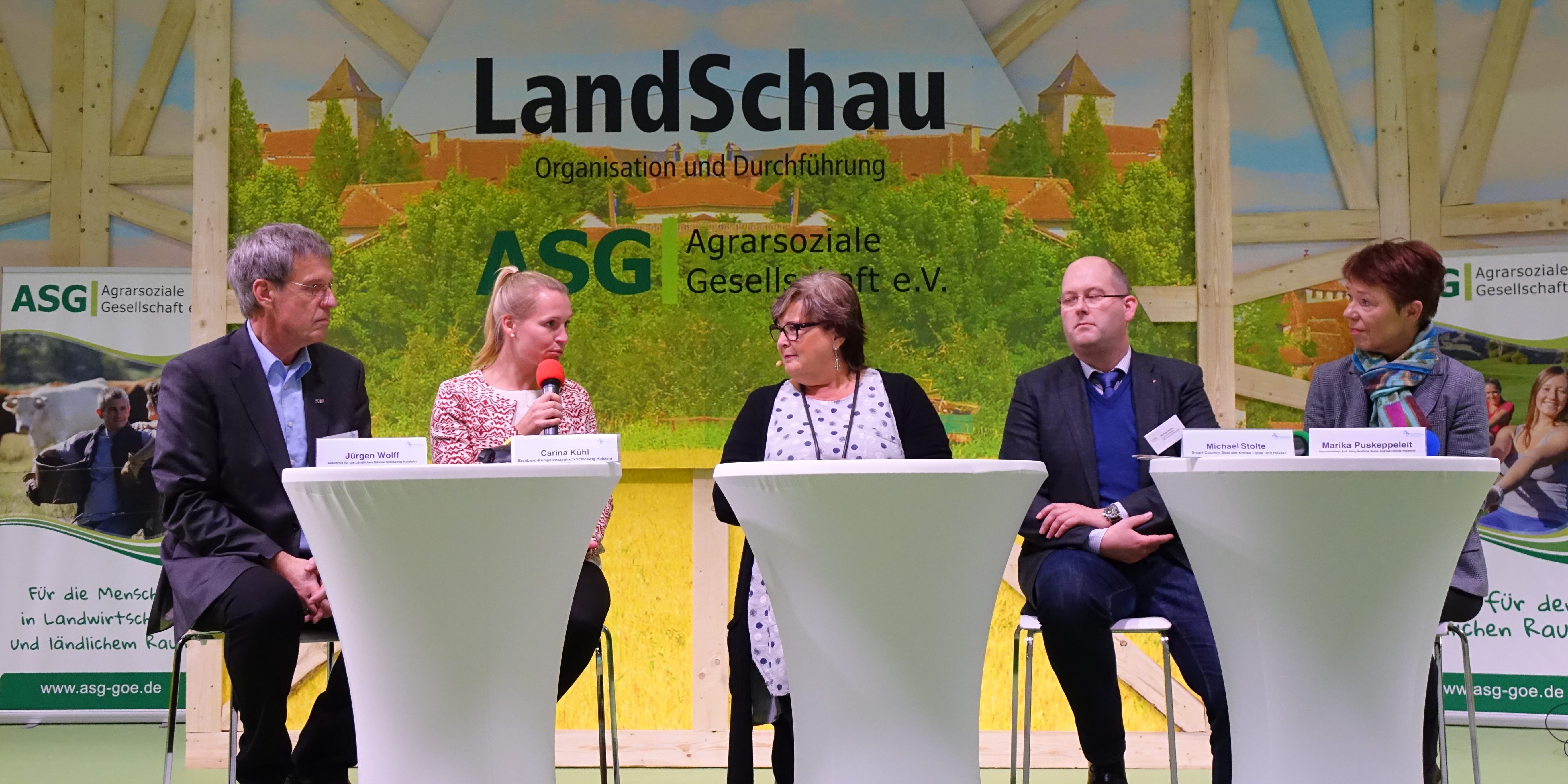 IGW 2019 Fünf Personen sitzen auf einer Bühne an Stehtischen und diskutieren. Im Hintergrund befindet sich ein Banner mit der Aufschrift "LandSchau" und "Agrarsoziale Gesellschaft e.V." Auf jeder Seite des Banners sind Werbebanner sichtbar.