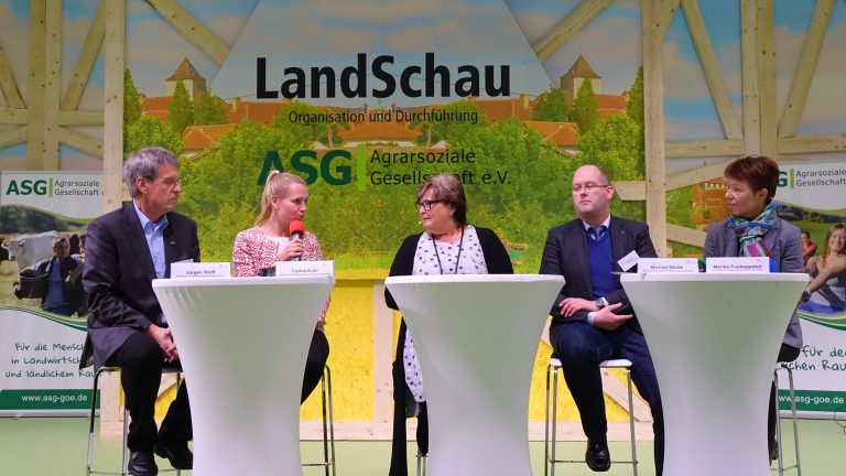 Fünf Personen sitzen auf einer Bühne an Stehtischen und diskutieren. Im Hintergrund befindet sich ein Banner mit der Aufschrift "LandSchau" und "Agrarsoziale Gesellschaft e.V." Auf jeder Seite des Banners sind Werbebanner sichtbar.