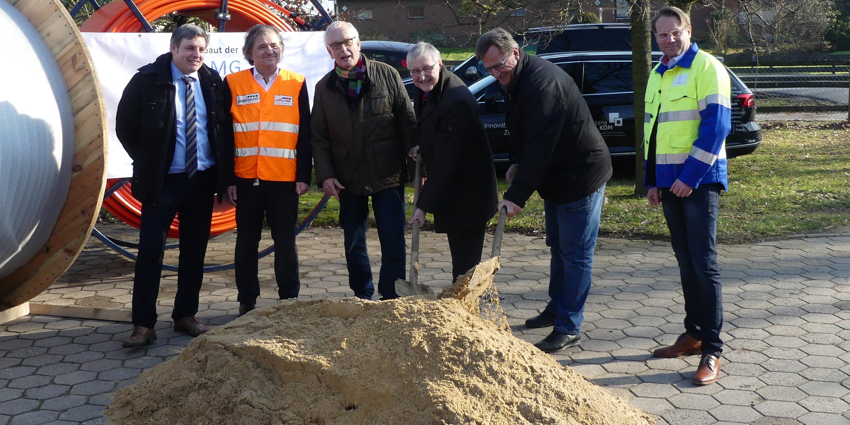 Spatenstich BZMG Sechs lächelnde Personen stehen auf einem gepflasterten Platz um einen Sandhaufen. Eine Person hält eine Schaufel darin. Im Hintergrund sind bunte Kabelrollen und Bäume zu sehen, einige Personen tragen farbige Westen.