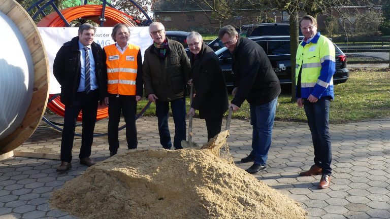 Sechs lächelnde Personen stehen auf einem gepflasterten Platz um einen Sandhaufen. Eine Person hält eine Schaufel darin. Im Hintergrund sind bunte Kabelrollen und Bäume zu sehen, einige Personen tragen farbige Westen.