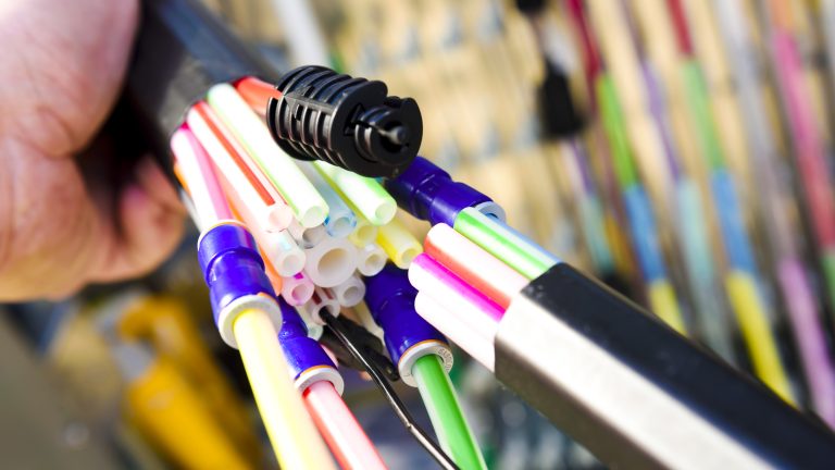 Nahaufnahme von einer Hand, die einen Strang bunter Glasfaserkabel hält. Die Kabel sind in einer Anordnung zusammengebunden und durch Verbindungsstücke miteinander verbunden. Im Hintergrund sind weitere Kabel unscharf zu sehen.
