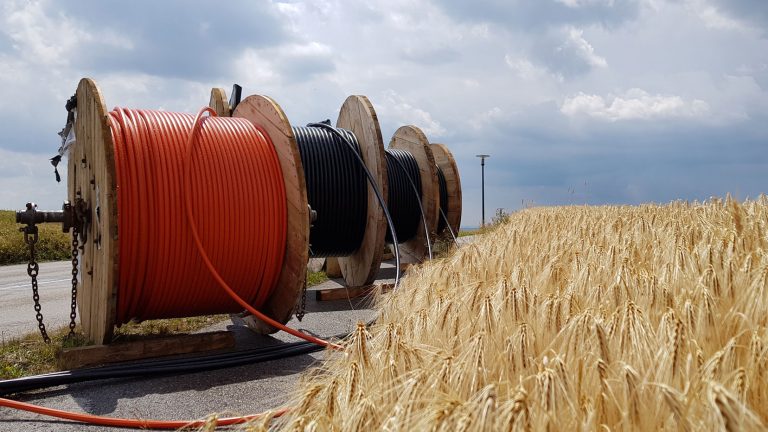 Drei große Kabeltrommeln mit jeweils einem orangefarbenen, schwarzen und blauen Kabel stehen nebeneinander auf einer asphaltierten Fläche neben einem reifen Getreidefeld, unter einem bewölkten Himmel.