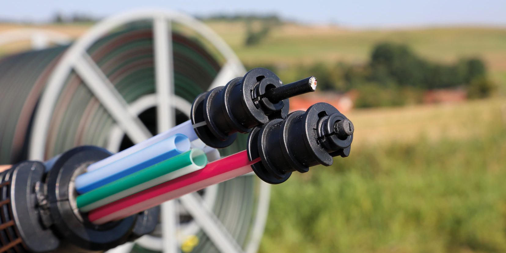 Eine Nahaufnahme von Glasfaserkabeln in einer Halterung, bestehend aus mehreren bunten, dünnen Röhren. Im Hintergrund verschwommen eine große Kabeltrommel und Landschaft mit Feldern und Bäumen.