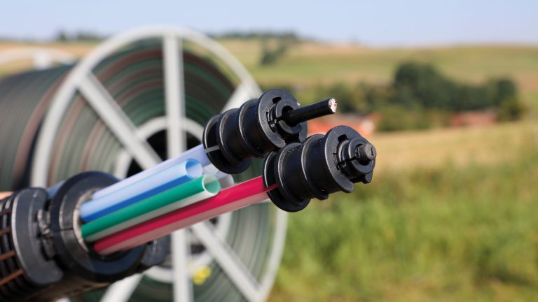 Eine Nahaufnahme von Glasfaserkabeln in einer Halterung, bestehend aus mehreren bunten, dünnen Röhren. Im Hintergrund verschwommen eine große Kabeltrommel und Landschaft mit Feldern und Bäumen.