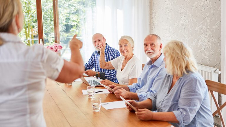 Eine Gruppe älterer Menschen sitzt an einem Holztisch in einem hellen Raum. Eine Person steht und spricht. Einige Personen sitzen mit Tablets, zwei heben die Hand. Im Hintergrund sind Fenster mit Blick ins Grüne.