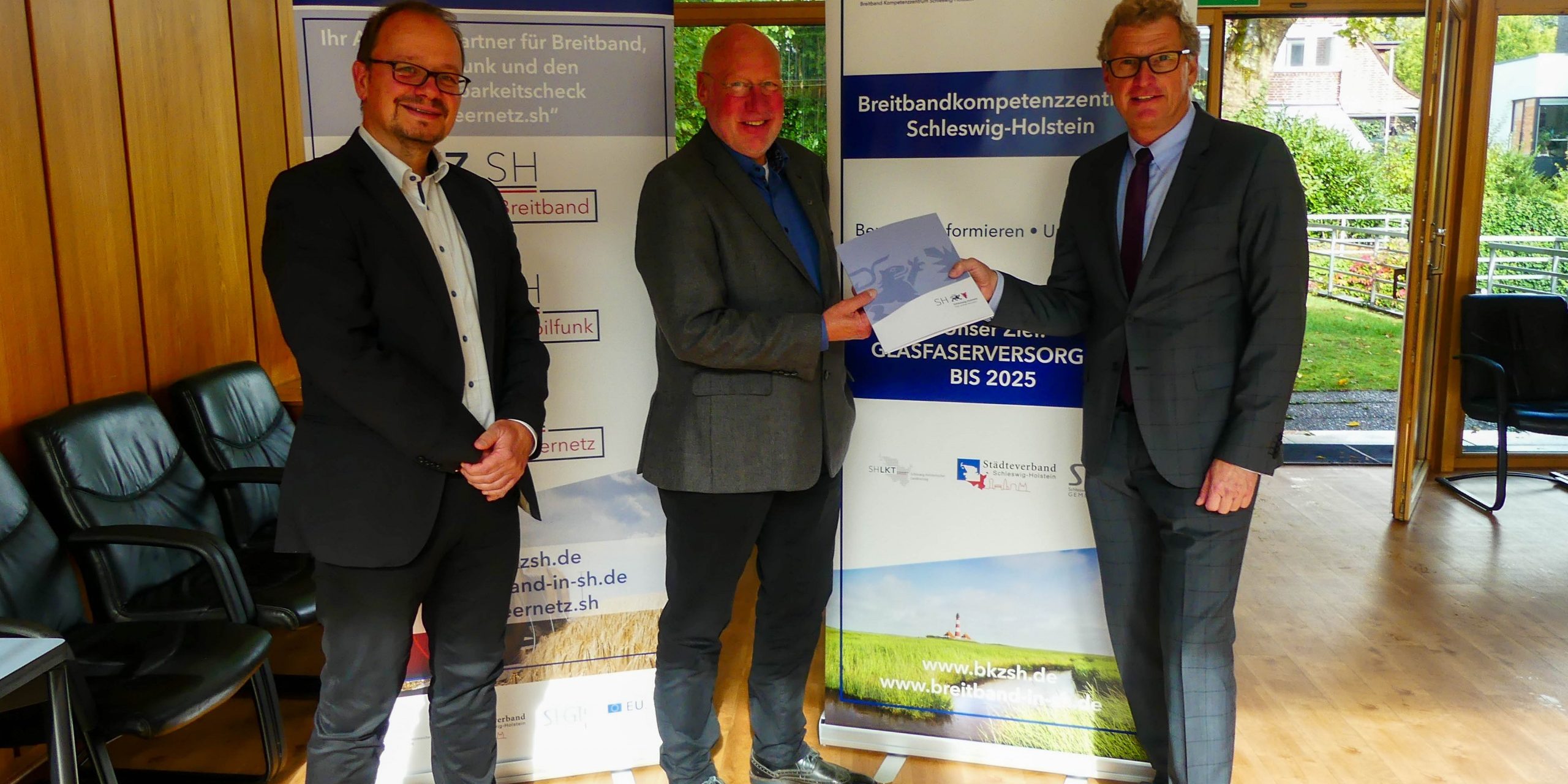 Drei Männer in Anzügen stehen in einem Raum mit Holzvertäfelung. Einer hält ein Dokument in der Hand. Im Hintergrund sind zwei Roll-Up-Banner mit Informationen über das Breitbandkompetenzzentrum Schleswig-Holstein.