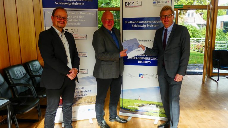Drei Männer in Anzügen stehen in einem Raum mit Holzvertäfelung. Einer hält ein Dokument in der Hand. Im Hintergrund sind zwei Roll-Up-Banner mit Informationen über das Breitbandkompetenzzentrum Schleswig-Holstein.