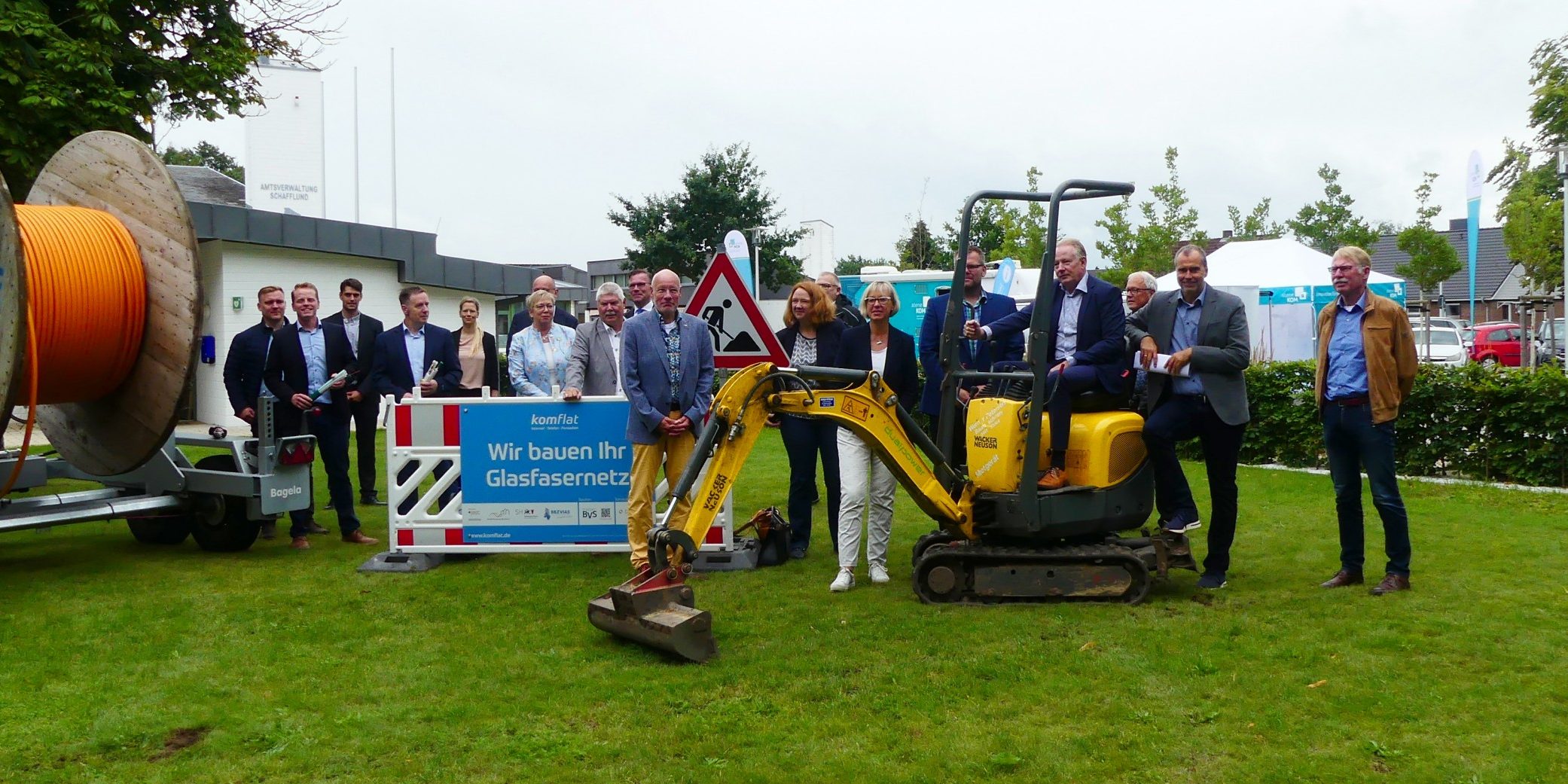 2021 Infotour Schafflund 13c Eine Gruppe von Menschen steht auf einer Wiese, neben einem kleinen Bagger und großen Kabelrollen. Schilder mit Text sind zu sehen. Im Hintergrund befinden sich Bäume und ein Gebäude.