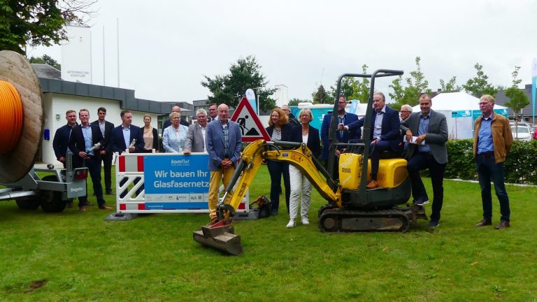 Eine Gruppe von Menschen steht auf einer Wiese, neben einem kleinen Bagger und großen Kabelrollen. Schilder mit Text sind zu sehen. Im Hintergrund befinden sich Bäume und ein Gebäude.