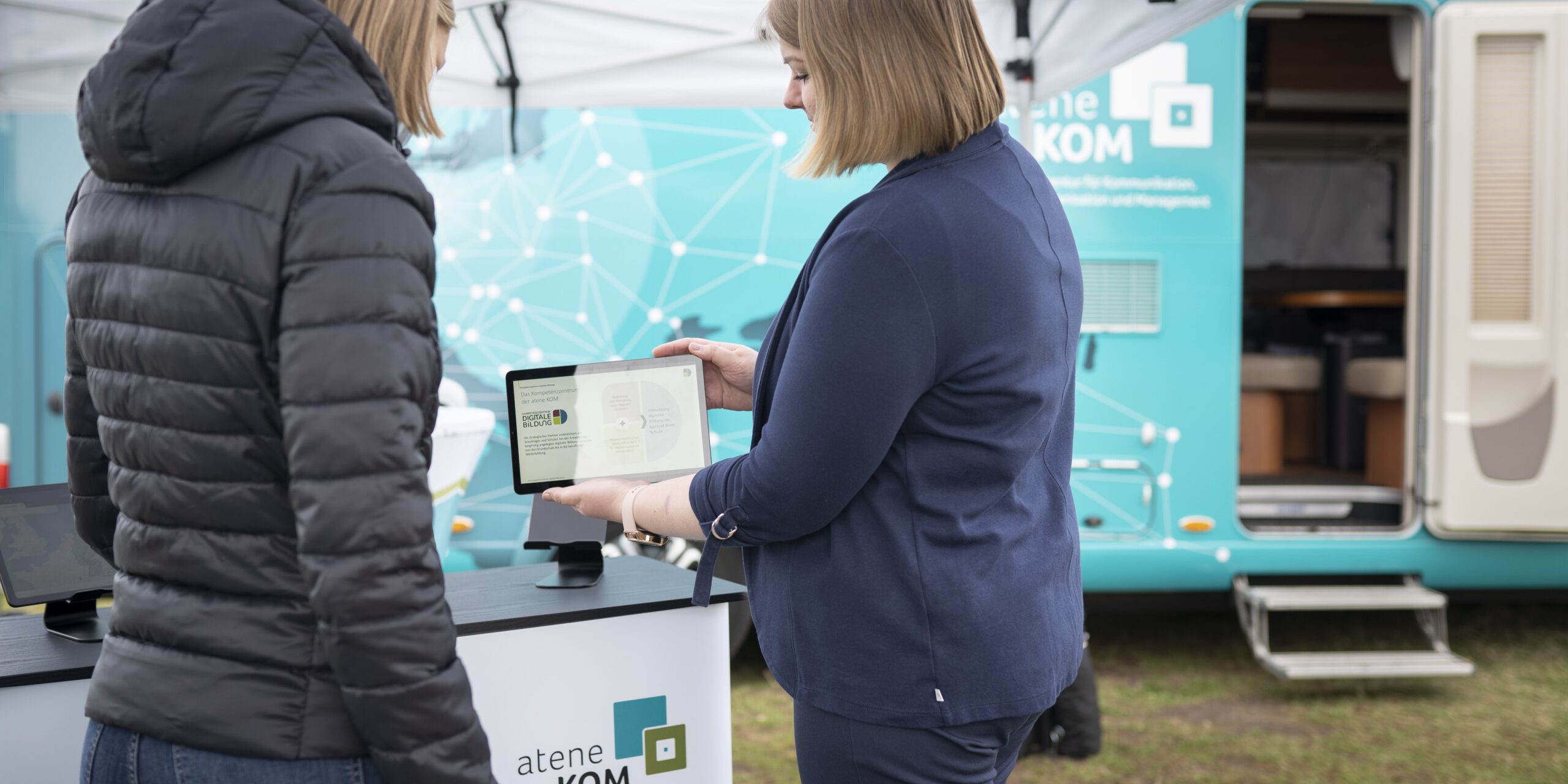 Roardshow „Zukunftsreise“ Zwei Personen stehen unter einem Zelt vor einem Informationstresen und betrachten ein Tablet. Im Hintergrund steht ein Anhänger mit aufgedrucktem Logo und Text.