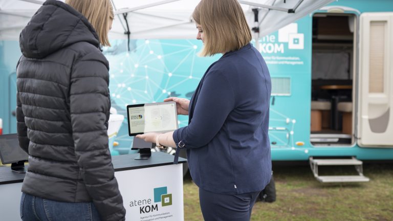 Zwei Personen stehen unter einem Zelt vor einem Informationstresen und betrachten ein Tablet. Im Hintergrund steht ein Anhänger mit aufgedrucktem Logo und Text.
