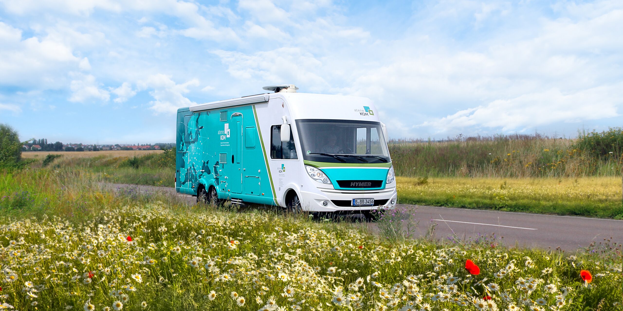 2021 Infotour Sylt 23 Ein weiß-blauer Bus fährt auf einer schmalen Landstraße durch eine grüne Landschaft mit Blumenwiese im Vordergrund und bewölktem Himmel im Hintergrund.