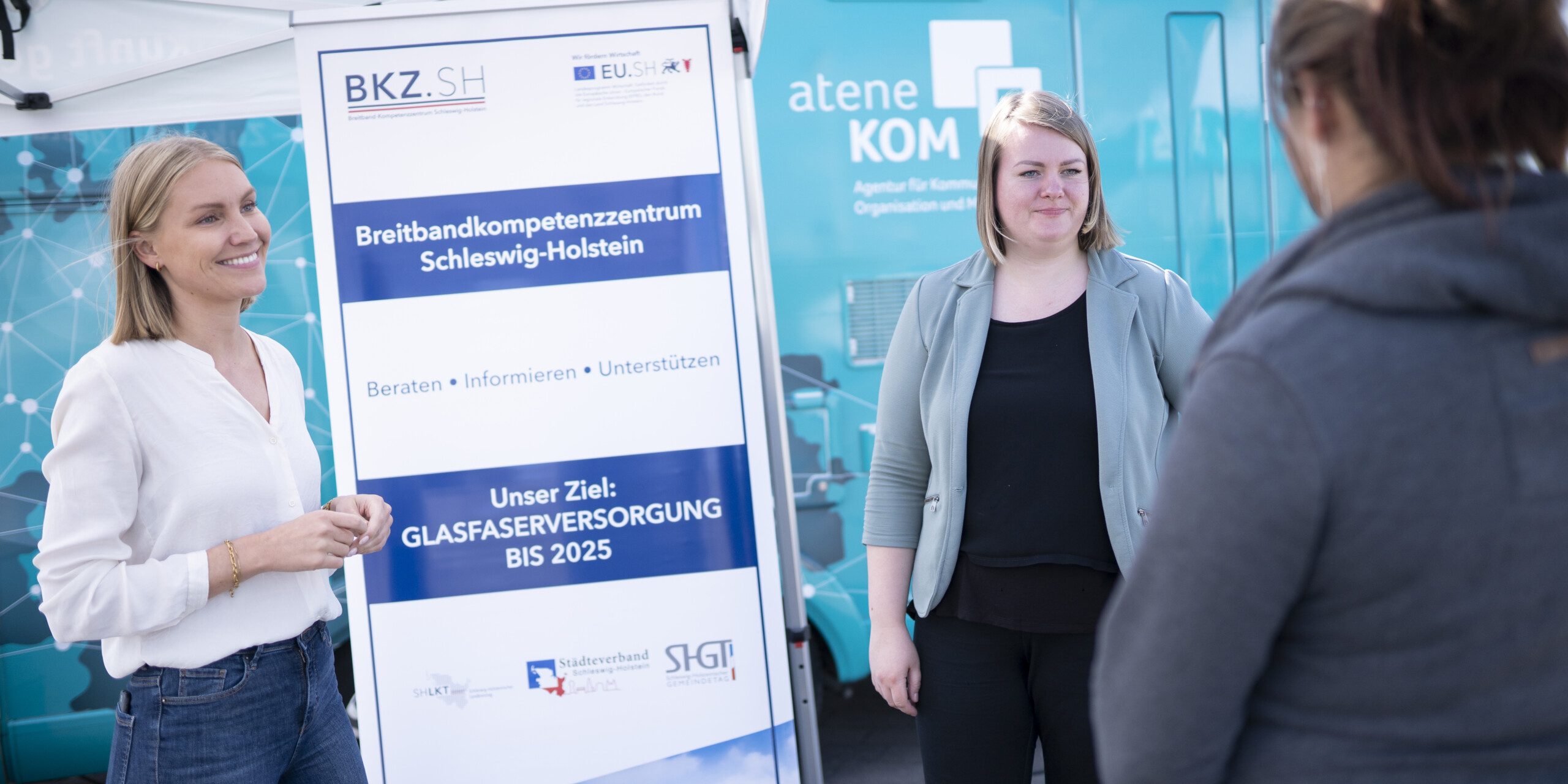 Roardshow „Zukunftsreise“ Zwei Personen stehen vor einem Informationsbanner zum Thema Breitbandversorgung. Eine dritte Person steht mit dem Rücken zur Kamera. Im Hintergrund ist ein blauer Infomobil-Anhänger zu sehen.