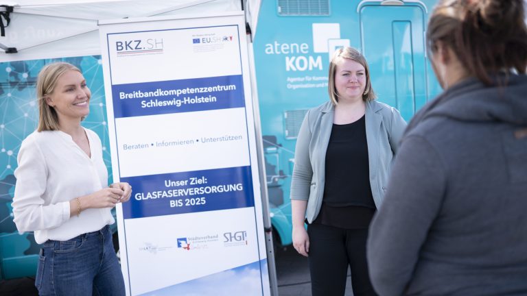 Zwei Personen stehen vor einem Informationsbanner zum Thema Breitbandversorgung. Eine dritte Person steht mit dem Rücken zur Kamera. Im Hintergrund ist ein blauer Infomobil-Anhänger zu sehen.