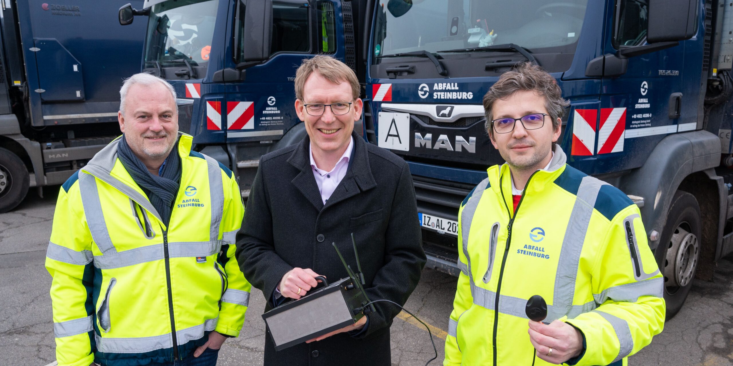 20230125 MobilfunkmessungKreisSteinburg Credit MichaelRuff Drei Männer stehen vor zwei blauen Müllfahrzeugen. Zwei tragen gelbe Sicherheitsjacken, der dritte einen dunklen Mantel. Einer hält ein Tablet. Alle lächeln in die Kamera. Die Fahrzeuge haben weiße und rote Streifen.