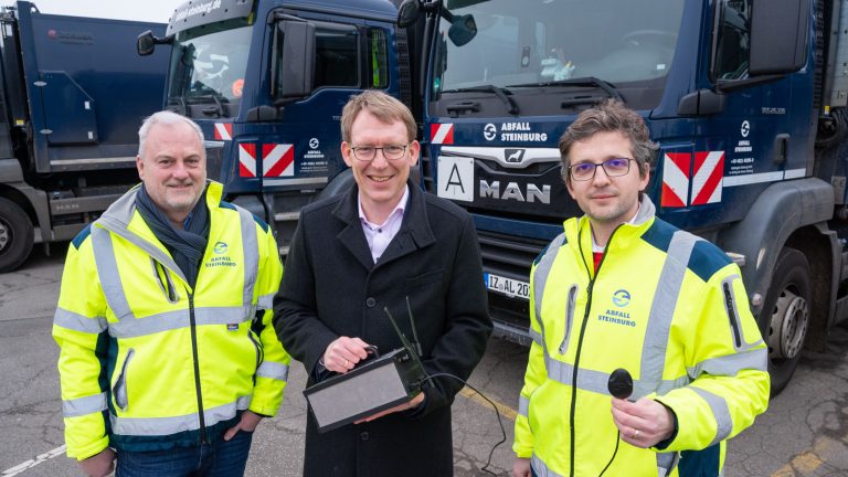 Drei Männer stehen vor zwei blauen Müllfahrzeugen. Zwei tragen gelbe Sicherheitsjacken, der dritte einen dunklen Mantel. Einer hält ein Tablet. Alle lächeln in die Kamera. Die Fahrzeuge haben weiße und rote Streifen.