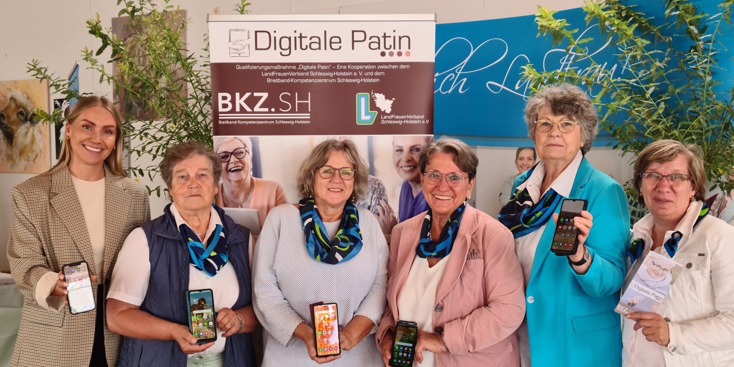 20230831 143236 Sechs Frauen stehen vor einem Banner mit der Aufschrift "Digitale Patin" und halten Smartphones. Sie lächeln in die Kamera. Im Hintergrund sind Pflanzen und weitere Banner zu sehen.