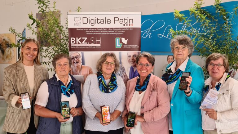 Sechs Frauen stehen vor einem Banner mit der Aufschrift "Digitale Patin" und halten Smartphones. Sie lächeln in die Kamera. Im Hintergrund sind Pflanzen und weitere Banner zu sehen.