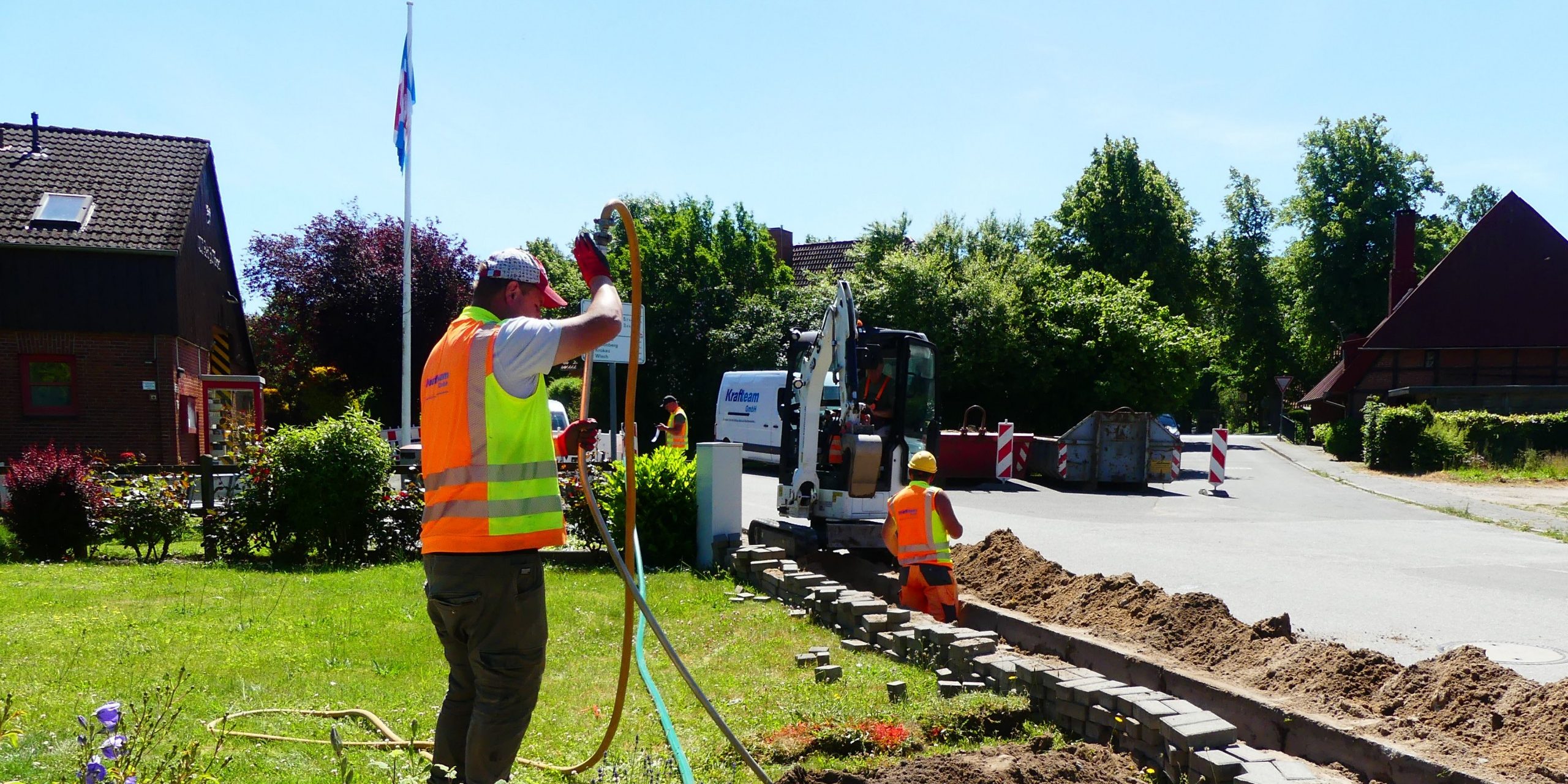 210616 Baustelle Barsbek Mehrere Bauarbeiter in Warnwesten führen Arbeiten an einer Straße durch. Ein Arbeiter bedient einen Bagger, während andere im Graben arbeiten. Im Hintergrund sind Häuser, Bäume und Baustellenabsperrungen zu sehen.