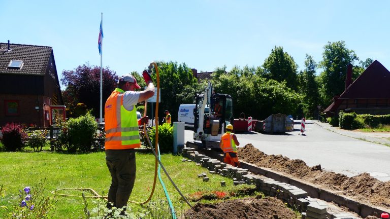 Mehrere Bauarbeiter in Warnwesten führen Arbeiten an einer Straße durch. Ein Arbeiter bedient einen Bagger, während andere im Graben arbeiten. Im Hintergrund sind Häuser, Bäume und Baustellenabsperrungen zu sehen.