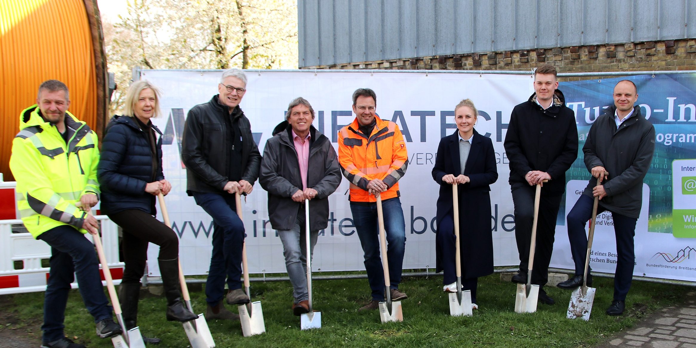 Eine Gruppe von acht Personen steht in einer Reihe vor einem Banner. Jede Person hält einen Spaten und gräbt symbolisch Erde um. Hinter ihnen sind ein Gebäude und einige Bäume zu sehen.