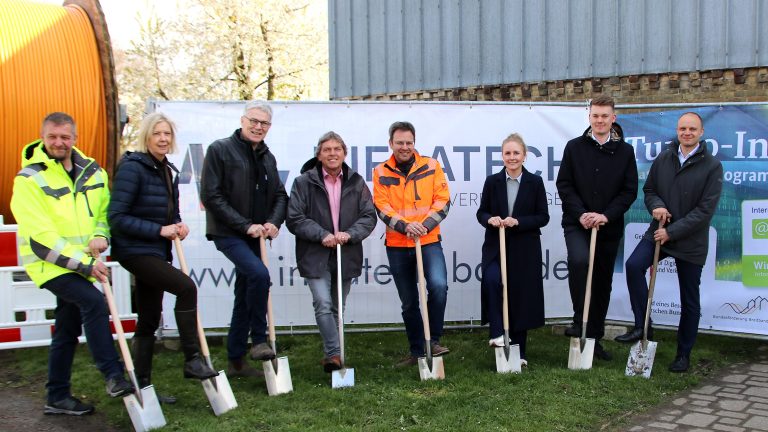 Eine Gruppe von acht Personen steht in einer Reihe vor einem Banner. Jede Person hält einen Spaten und gräbt symbolisch Erde um. Hinter ihnen sind ein Gebäude und einige Bäume zu sehen.