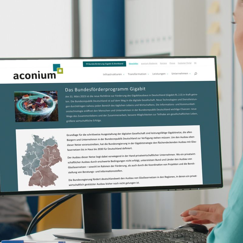 Eine Person sitzt an einem Schreibtisch und schaut auf einen Computerbildschirm, der eine Webseite mit dem Titel "Das Bundesförderprogramm Gigabit" anzeigt. Im Hintergrund arbeitet eine weitere Person an einem Tisch.