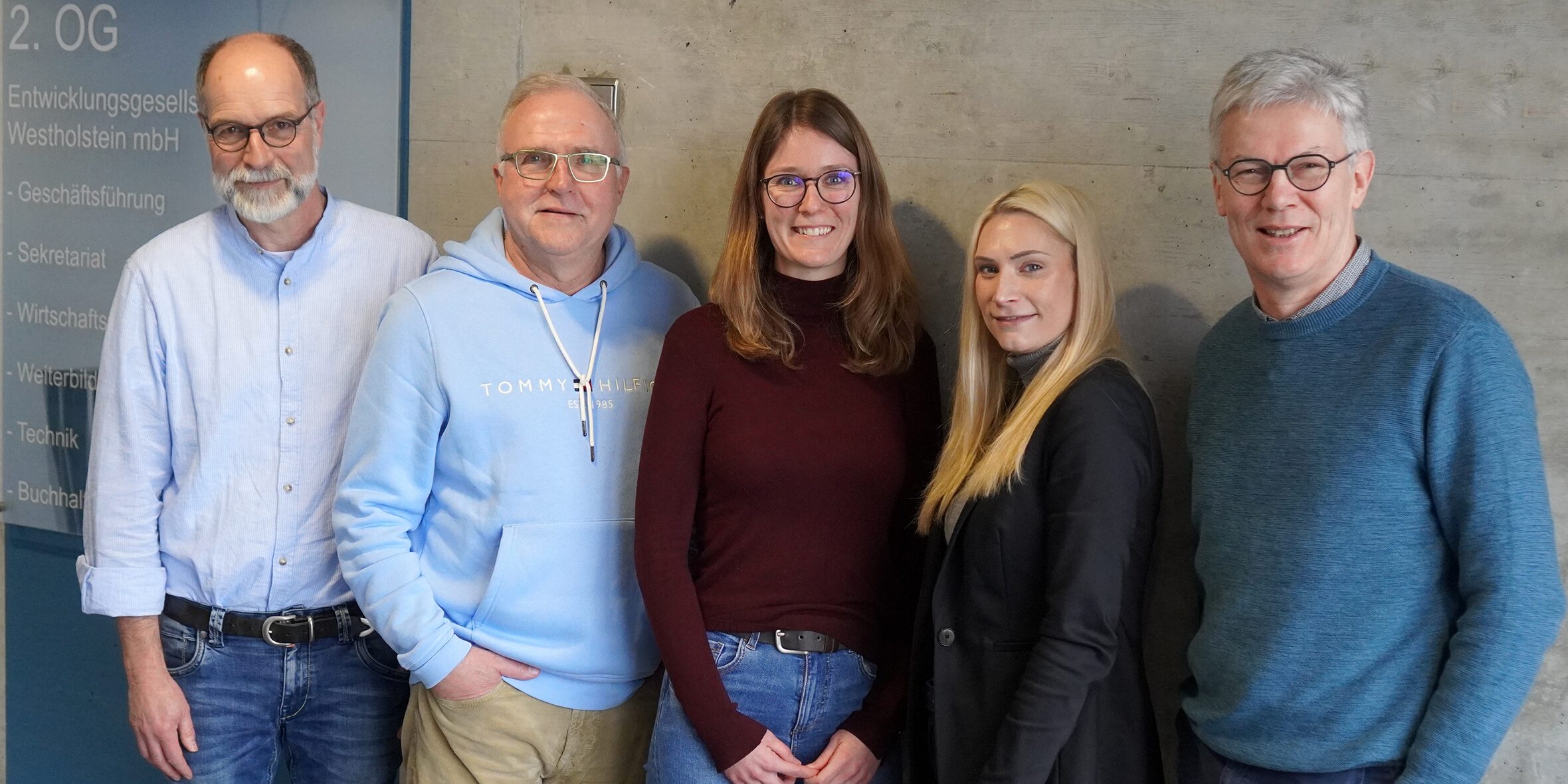 250116 Egw ZVBS Fünf Personen stehen lächelnd nebeneinander vor einer Betonwand. Zwei Männer tragen Brillen. Alle sind lässig gekleidet. Links ist ein Schild mit Aufschrift zu sehen.