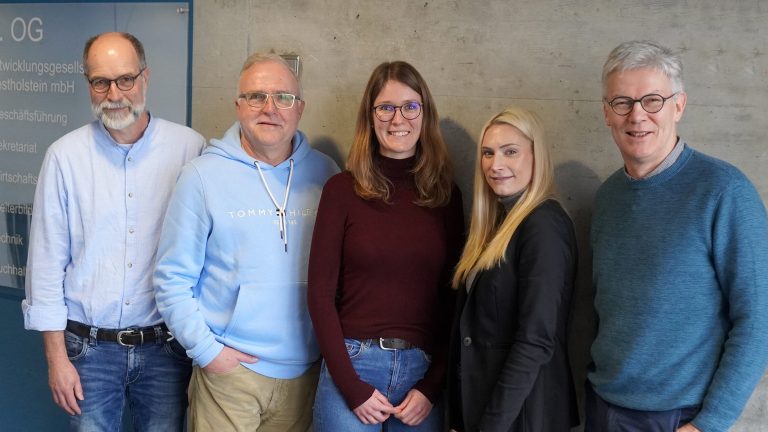 Fünf Personen stehen lächelnd nebeneinander vor einer Betonwand. Zwei Männer tragen Brillen. Alle sind lässig gekleidet. Links ist ein Schild mit Aufschrift zu sehen.