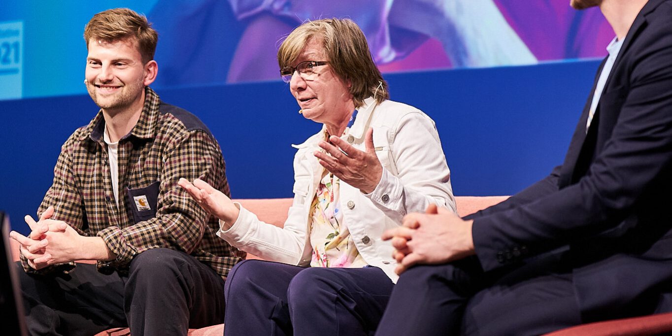Drei Personen sitzen auf einer Bühne auf Sofas. Sie diskutieren, während sie Mikrofone tragen. Eine Person gestikuliert mit den Händen. Im Hintergrund ist ein blaues Banner. Alle drei tragen legere Kleidung und Turnschuhe.