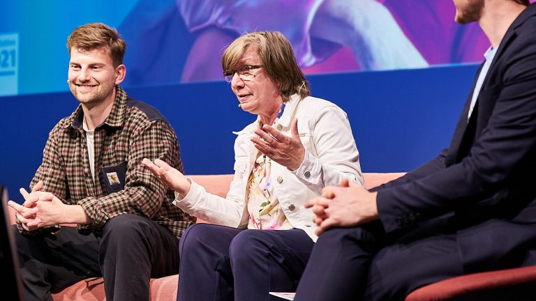 Drei Personen sitzen auf einer Bühne auf Sofas. Sie diskutieren, während sie Mikrofone tragen. Eine Person gestikuliert mit den Händen. Im Hintergrund ist ein blaues Banner. Alle drei tragen legere Kleidung und Turnschuhe.