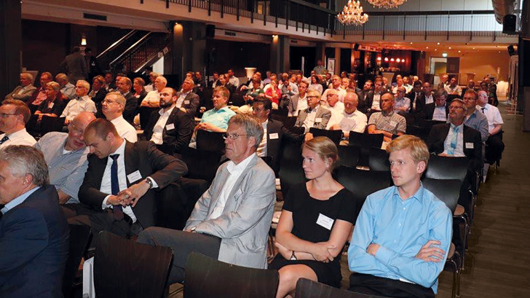 Ein voll besetzter Konferenzsaal mit Menschen unterschiedlichen Alters und Geschlechts, die aufmerksam einem Vortrag folgen. Der Raum ist gut beleuchtet und verfügt über Kronleuchter an der Decke.