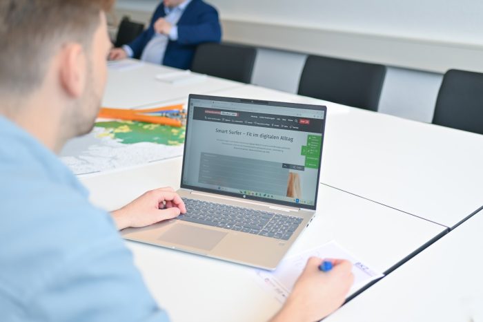 Eine Person sitzt an einem weißen Tisch und benutzt einen Laptop. Auf dem Bildschirm ist eine Website mit dem Titel "Smart Surfer – fit im digitalen Alltag" zu sehen. Im Hintergrund sitzt eine weitere Person.