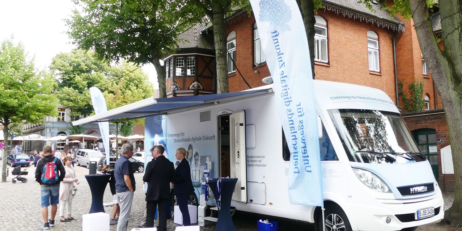 BKZSH Inseltour Fehmarn Markt 2018 Ein weißer Infomobil steht auf einem gepflasterten Platz, umgeben von vier Personen. Es hat eine Markise und Fahnen mit Aufschrift. Im Hintergrund sind Bäume und ein Backsteingebäude zu sehen.
