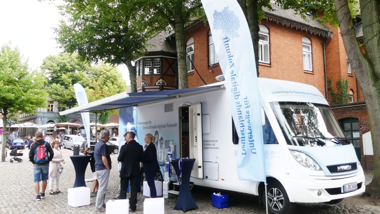 Ein weißer Infomobil steht auf einem gepflasterten Platz, umgeben von vier Personen. Es hat eine Markise und Fahnen mit Aufschrift. Im Hintergrund sind Bäume und ein Backsteingebäude zu sehen.