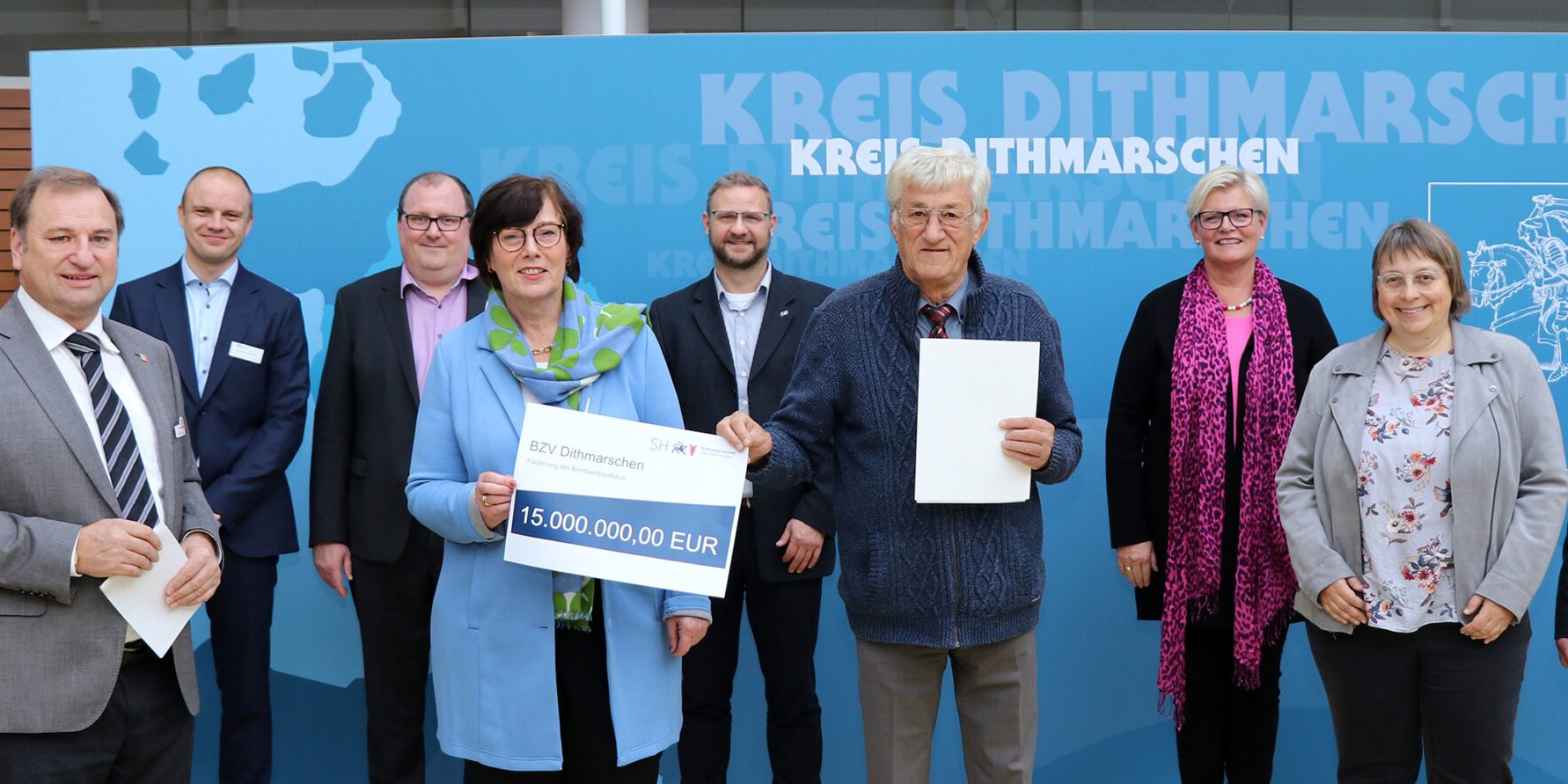 BZVD 1901 Zugeschnitten Eine Gruppe von Menschen steht vor einer blauen Wand mit der Aufschrift "Kreis Dithmarschen". Eine Person hält einen symbolischen Scheck über 1,5 Millionen Euro. Alle blicken in die Kamera und lächeln.