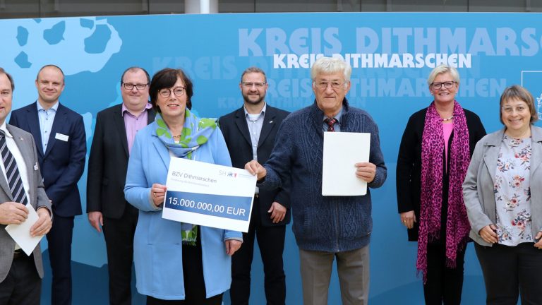 Eine Gruppe von Menschen steht vor einer blauen Wand mit der Aufschrift "Kreis Dithmarschen". Eine Person hält einen symbolischen Scheck über 1,5 Millionen Euro. Alle blicken in die Kamera und lächeln.
