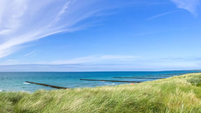 Wellen schlagen gegen Holzbuhnen im klaren, blauen Meer. Im Vordergrund bewegen sich hohe Gräser im Wind. Der Himmel ist weit und blau mit leichten Wolken, die darüber ziehen.