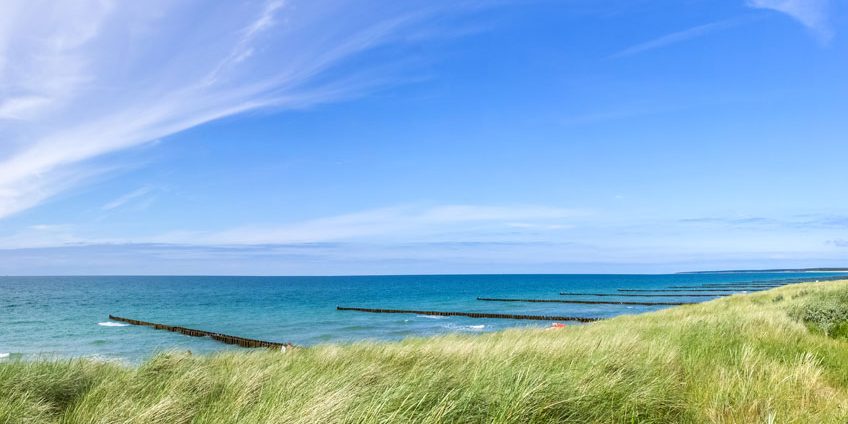 Wellen schlagen gegen Holzbuhnen im klaren, blauen Meer. Im Vordergrund bewegen sich hohe Gräser im Wind. Der Himmel ist weit und blau mit leichten Wolken, die darüber ziehen.
