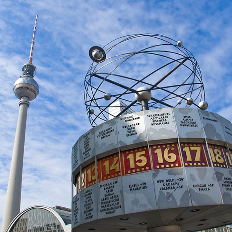 Der Berliner Fernsehturm und die Weltzeituhr auf dem Alexanderplatz. Die Uhr zeigt verschiedene Städte und Zeiten an. Der Himmel ist leicht bewölkt. Die Perspektive ist von unten nach oben.