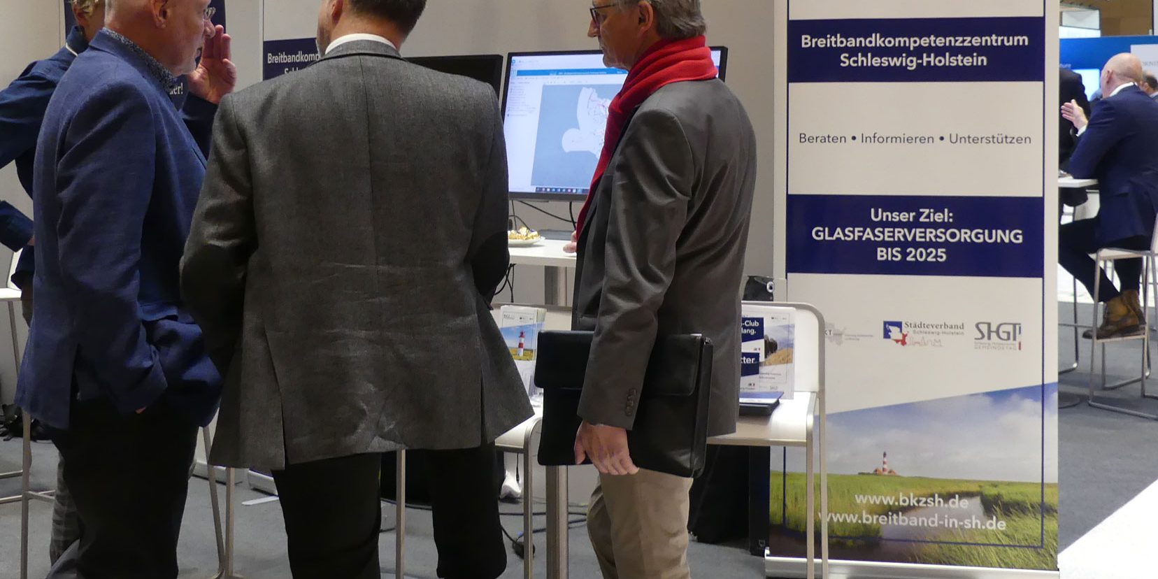 Drei Männer in Anzügen stehen vor einem Informationsstand mit Bannern und Plakaten. Auf einem Banner steht: "Breitbandkompetenzzentrum Schleswig-Holstein". Ein Mann trägt einen roten Schal. Auf dem Tisch sind Computer zu sehen.