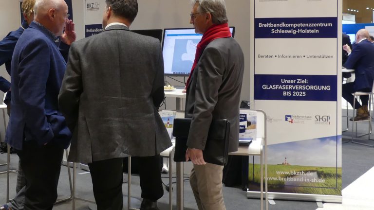 Drei Männer in Anzügen stehen vor einem Informationsstand mit Bannern und Plakaten. Auf einem Banner steht: "Breitbandkompetenzzentrum Schleswig-Holstein". Ein Mann trägt einen roten Schal. Auf dem Tisch sind Computer zu sehen.