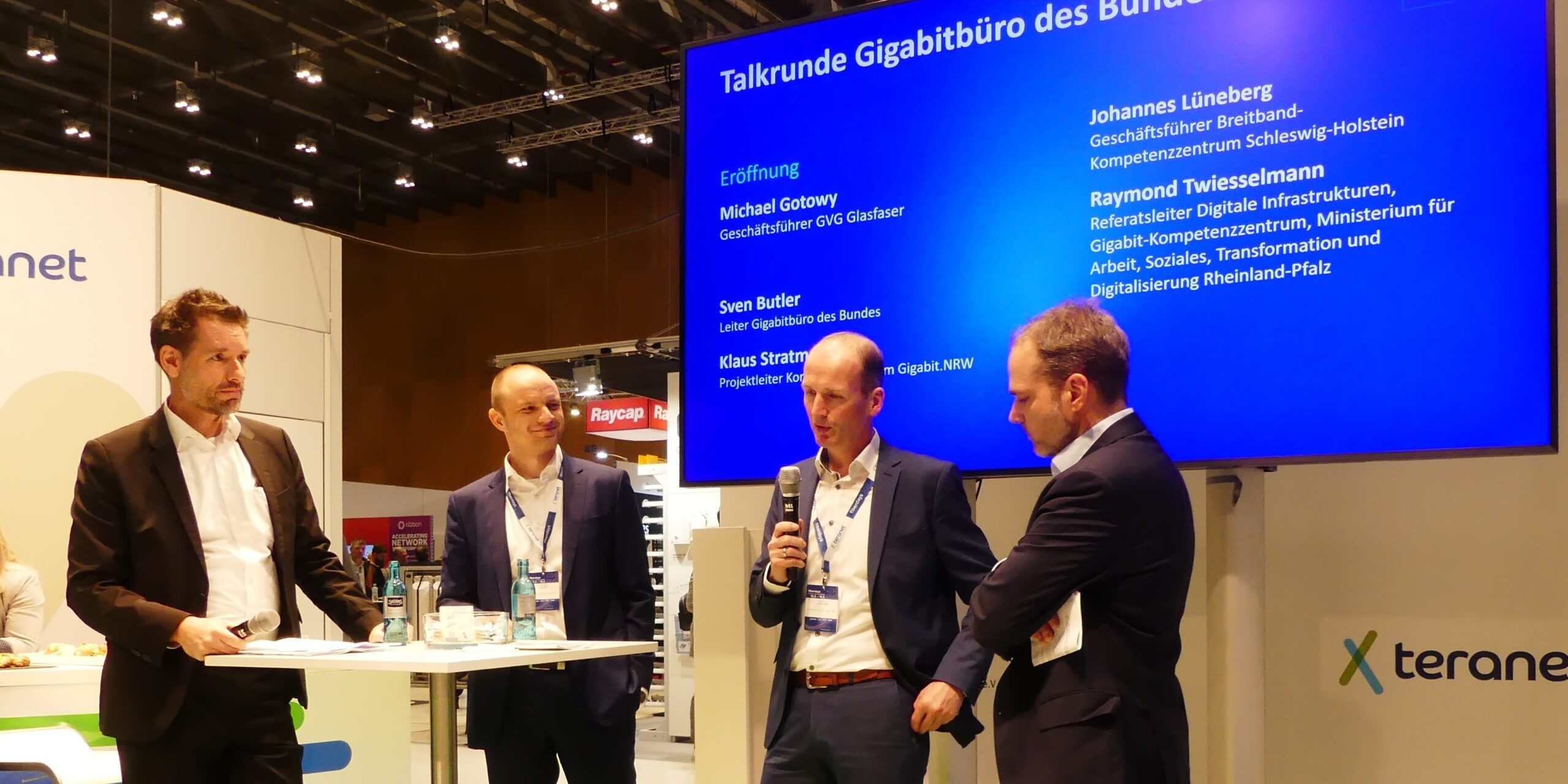 FiberdaysTalkrunde Zuschnitt Homepage Vier Männer in Anzügen stehen auf einer Bühne vor einem großen Bildschirm, auf dem "Talkrunde Gigabüro des Bundes" steht. Sie halten Mikrofone und scheinen in ein Gespräch vertieft zu sein. Im Hintergrund sind Ausstellungsstände zu sehen.