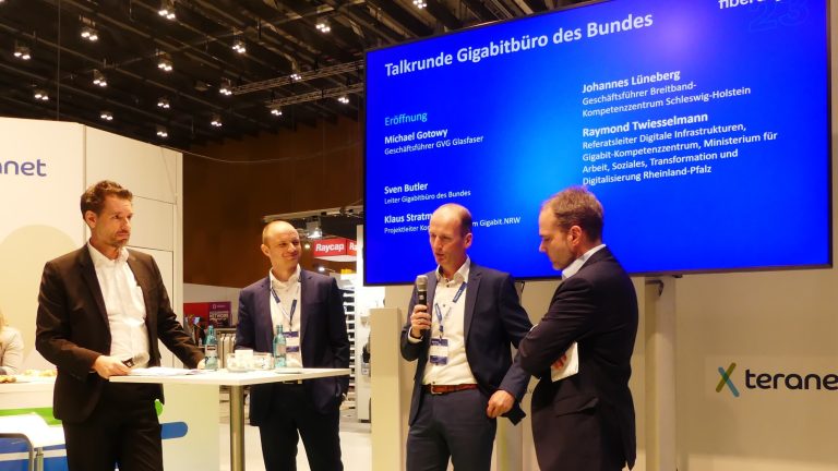 Vier Männer in Anzügen stehen auf einer Bühne vor einem großen Bildschirm, auf dem "Talkrunde Gigabüro des Bundes" steht. Sie halten Mikrofone und scheinen in ein Gespräch vertieft zu sein. Im Hintergrund sind Ausstellungsstände zu sehen.