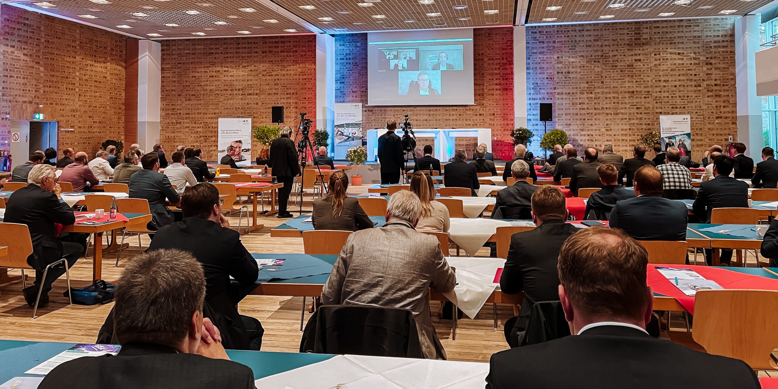 In einem Konferenzraum sitzen viele Personen an Tischen, die auf eine Bühne mit einer Leinwand gerichtet sind. Auf der Leinwand sind vier Personen in einem Videocall zu sehen. Die Bühne ist mit Pflanzen dekoriert.