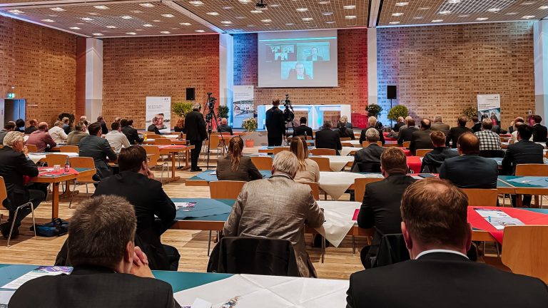 In einem Konferenzraum sitzen viele Personen an Tischen, die auf eine Bühne mit einer Leinwand gerichtet sind. Auf der Leinwand sind vier Personen in einem Videocall zu sehen. Die Bühne ist mit Pflanzen dekoriert.