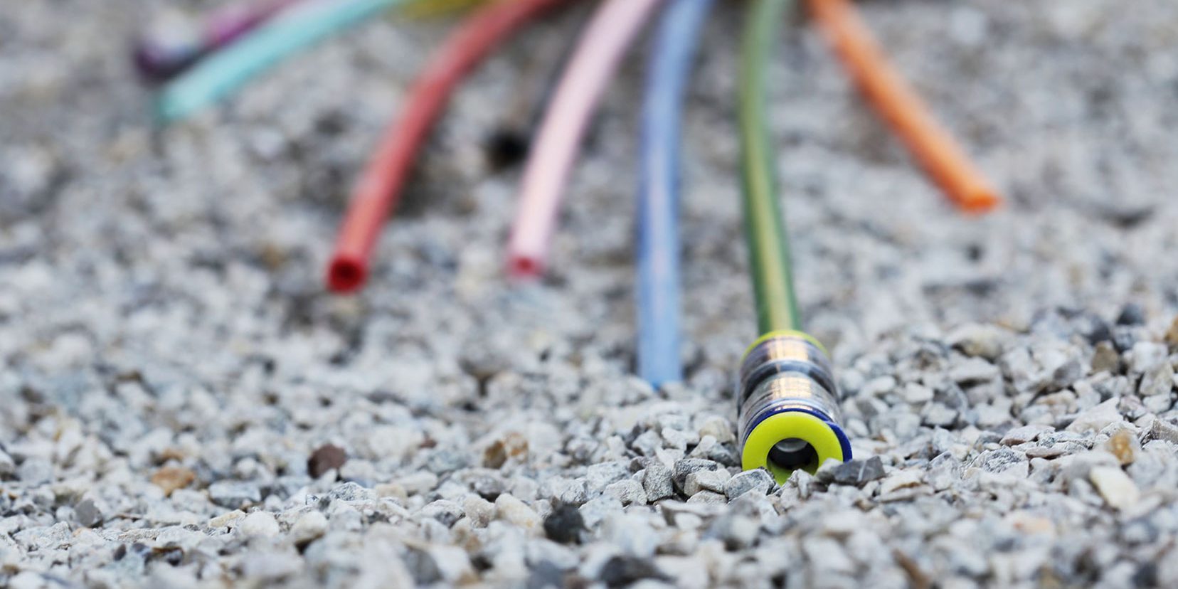 Glasfaserkabel Stein Header Bunte Kabel oder Rohre liegen auf einem Kiesbett verstreut. Ein Kabel ist im Vordergrund scharf fokussiert, während der Hintergrund unscharf erscheint.