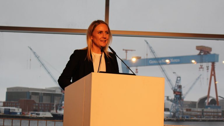 Eine Person steht lächelnd hinter einem Rednerpult in einem Gebäude mit großen Fenstern. Im Hintergrund sind ein Hafenkran und Schiffe zu sehen. Das Bild ist in warmem Licht gehalten.